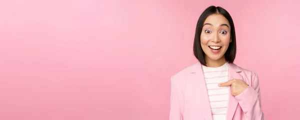 Portrait de jeune femme d'affaires asiatique avec une expression du visage surprise et excitée, pointant du doigt vers elle-même, debout en costume sur fond rose — Photo