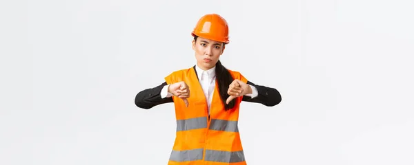 Розчарований і засмучений азіатський головний інженер оглядає погану роботу, показуючи великі пальці вниз розчаровані, носити безпечний шолом і скаржитися на слабку жахливу роботу архітектора, білий фон — стокове фото