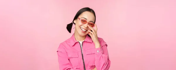Крупный план портрета стильной азиатской женщины в солнечных очках, улыбающейся, выглядящей польщенной, смеющейся кокетливой, стоящей на розовом фоне — стоковое фото