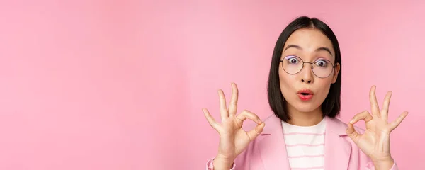 Close up portrait of business woman looks impressed and shows okay sign in approval, recommending company. Young corporate lady in glasses shows ok gesture, pink background — Stock Photo, Image