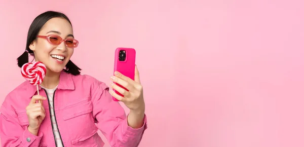 Porträtt av snygg, glad asiatisk flicka som tar selfie med godis, godis, godis godis och leende, ta foto med mobilapp, står över rosa bakgrund — Stockfoto