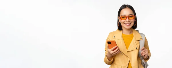 Snygg ung asiatisk kvinna turist, resenär med ryggsäck och smartphone ler mot kameran, poserar mot vit bakgrund — Stockfoto