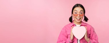 Güzel Asyalı kız mutlu gülümsüyor, kalp hediye kutusunu gösteriyor ve kameraya heyecanlı bakıyor, pembe romantik arka planda duruyor.
