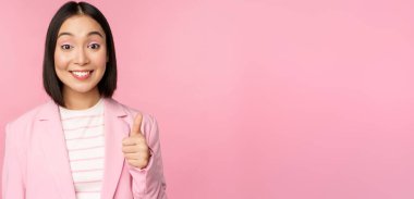 Asyalı iş kadını portresi tatmin olmuş gülümsüyor, başparmaklarını gösteriyor, övüyor ve onaylıyor, pembe arka planda takım elbise içinde duruyor.