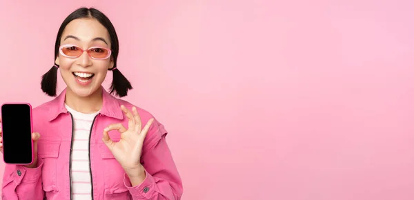 Entusiasta joven mujer asiática mostrando bien, ok signo, sonriendo complacido, pantalla del teléfono móvil, aplicación de teléfono inteligente, de pie sobre fondo rosa — Foto de Stock