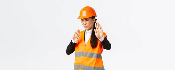 Nespokojený a zdráhavý asijský architekt v bezpečnostní helmě šklebící se z averze, ukazující stop gesto, odmítající nebo odmítající odporný nechutný plán, stojící zklamané bílé pozadí — Stock fotografie