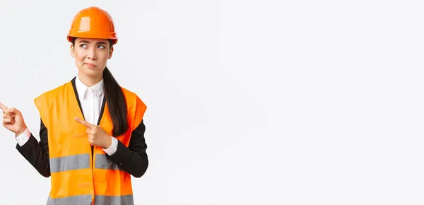 Budownictwo, budownictwo i koncepcja przemysłowa. niezdecydowana azjatycka kobieta architekt jest niepewny, nosząc odblaskową odzież i helmer, uśmiech jak wskazując i patrząc w lewym górnym rogu zastanawiające — Zdjęcie stockowe