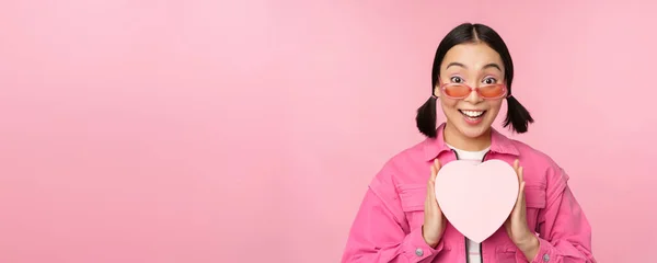 Όμορφη ασιατική κοπέλα χαμογελά ευτυχισμένη, δείχνει καρδιά κουτί δώρου και αναζητούν ενθουσιασμένοι στην κάμερα, στέκεται πάνω από ροζ ρομαντικό φόντο — Φωτογραφία Αρχείου
