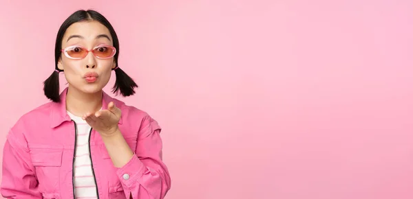 Elegante ragazza k-pop in occhiali da sole, donna asiatica che soffia aria bacio alla macchina fotografica, labbra pucker, gesto mwah, in piedi su sfondo studio rosa — Foto Stock