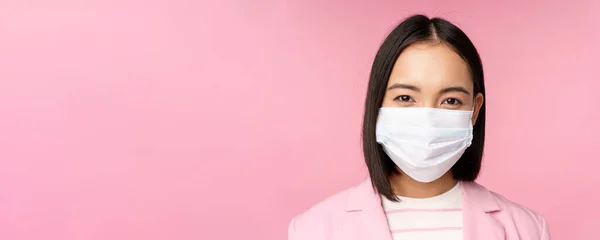 Närbild porträtt av japanska affärskvinna i medicinsk ansiktsmask, kostym, tittar på kameran, står över rosa bakgrund — Stockfoto