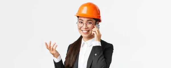 Close-up van Aziatische zakenvrouw beheren onderneming, ingenieur in veiligheid helm en pak met telefoon gesprek, bellen beleggers, glimlachen tijdens het praten over smartphone, witte achtergrond — Stockfoto