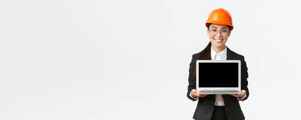 Lächelnde professionelle asiatische Ingenieurin stellt Investoren oder Bauherren Bauplan vor, steht in Schutzhelm und Anzug und zeigt Laptop-Bildschirm mit zufriedenem Lächeln, weißer Hintergrund — Stockfoto