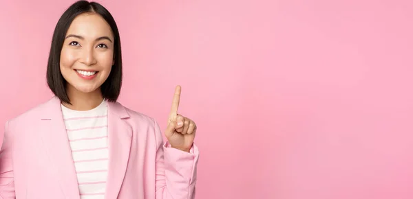 Uśmiechnięta koreańska bizneswoman, wskazująca palcem w górę, pokazująca reklamę, baner lub logo na górze, stojąca w garniturze na różowym tle — Zdjęcie stockowe
