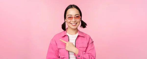 Retrato de chica asiática sonriente en traje elegante, gafas de sol, señalando el dedo a la izquierda, mostrando publicidad, pancarta, de pie sobre fondo rosa —  Fotos de Stock