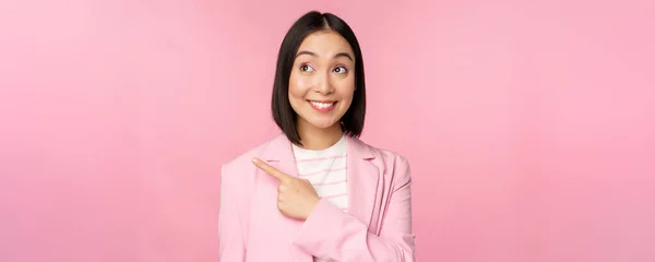 Enthousiaste jeune femme d'affaires asiatique, employé de bureau pointant du doigt vers la gauche, regardant bannière, publicité avec sourire heureux, montrant la publicité, fond rose — Photo