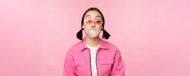 Havalı Asyalı kız sakız şişiriyor, sakız çiğniyor, güneş gözlüğü takıyor, pembe arka planda poz veriyor.