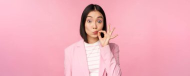 Asyalı kurumsal bir kadının portresi, ağzı mühürlü, dudakları kapalı anahtar hareketi, söz ver sır tut, pembe arka planda duran takım elbiseli.