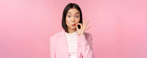 Retrato de mujer corporativa asiática mostrando sello bucal, cerrar los labios en el gesto clave, promesa mantener en secreto, de pie sobre fondo rosa en traje —  Fotos de Stock