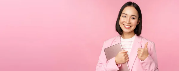 Retrato de la mujer corporativa, chica en la oficina en traje de negocios, sosteniendo tableta digital, mostrando los pulgares hacia arriba, recomendando la empresa, de pie sobre el fondo rosa —  Fotos de Stock