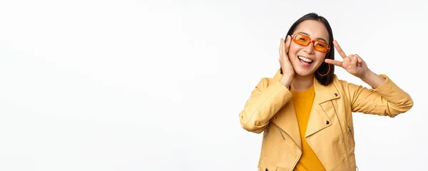 Retrato de chica moderna asiática con estilo, con gafas de sol y chaqueta amarilla, mostrando paz, gesto de signo v, de pie sobre fondo blanco, cara sonriente feliz — Foto de Stock