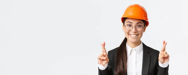 Close-up de animado esperançoso engenheiro asiático feminino, gerente de construção em capacete de segurança e terno de negócios implorando por bom gosto, dedos cruzados boa sorte e mordendo lábio tentador, fundo branco — Fotografia de Stock