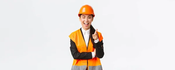Mosolygó professzionális ázsiai női építőipari vezető biztonsági sisak és fényvisszaverő kabát invitáló check-out új épület, kacsintás és ujjal mutogató kamera, bemutató ügyfél legjobb ingatlan — Stock Fotó