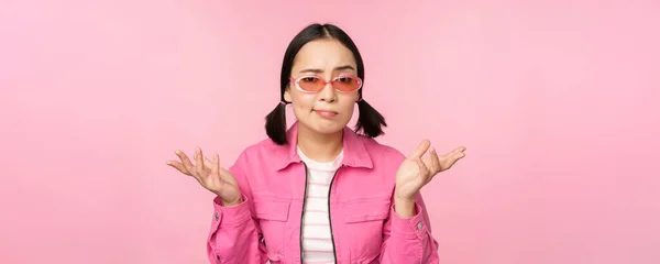 Primer plano retrato de asiático chica buscando confundido, encogiéndose de hombros desconcertado y mirando a cámara, usando gafas de sol, de pie sobre rosa fondo —  Fotos de Stock