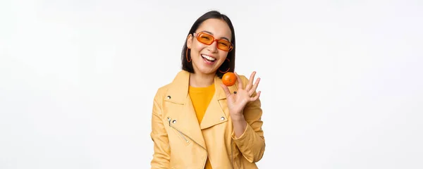 Menina asiática bonita em óculos de sol mostrando tangerina e sorrindo, olhando feliz, posando em amarelo contra o fundo do estúdio — Fotografia de Stock