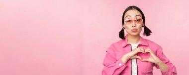 Güneş gözlüklü sevimli Koreli kadın model, kalbi gösteriyor, dikkat işareti, jestini seviyorum, pembe arka planda duruyor.