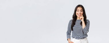 Yaşam tarzı, insanların duyguları ve gelişigüzel kavramlar. Utangaç, genç Asyalı kızın bir fikri var, bir şey söylemek istiyor ama tereddüt ediyor, parmağını ısırıyor ve garip bir şekilde gülümsüyor, beyaz arka planda duruyor.