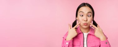 Gamzelerini gösteren genç Asyalı kızın yakın portresi, yanaklarını dürtüyor, komik suratlar yapıyor, pembe arka planda duruyor.