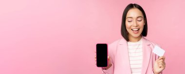 Gülümseyen Koreli iş kadını, cep telefonu ekranı, kredi kartı, online bankacılık başvuru arayüzü, pembe arka plan