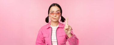 Güneş gözlüklü, ciddi, stil sahibi Asyalı bir kızın görüntüsü, durduğunu gösteriyor, jesti yasaklıyor, tabu işareti, hayır diyor, pembe arka planda duruyor.