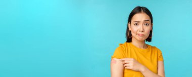 Covid-19 'dan aşı kampanyası. Üzgün ve hayal kırıklığına uğramış Asyalı kadın omzunu bandajlı ve asık suratlı, mavi arka planda dikiliyor.