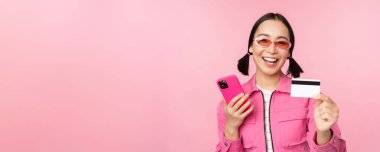 Modern, güzel Asyalı kız, cep telefonu, kredi kartı, online alışveriş, akıllı telefon ile ödeme, pembe arka planda durmak