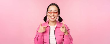 Güneş gözlüklü modern Asyalı kızın yakın portresi gülümsüyor, kamerayı işaret ediyor, seni övüyor, davet ediyor ya da iltifat ediyor, pembe arka planda duruyor.