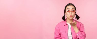 Güneş gözlüklü şık k-pop kızı, kameraya hava öpücüğü veren Asyalı kadın, büzüşmüş dudaklar, mwah jest, pembe stüdyo arka planında duran