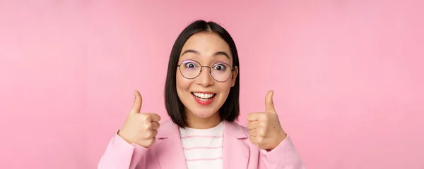Wspaniale, gratulacje. Twarz podekscytowanej azjatyckiej bizneswoman w okularach, uśmiechnięty zadowolony, pokazując kciuki w górę w aprobacie, stojąc na różowym tle — Zdjęcie stockowe
