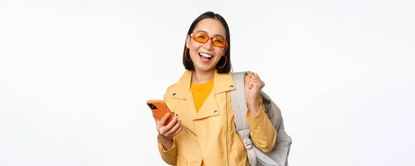 Joven mujer asiática viajero, chica turista en gafas de sol, la celebración de la mochila y el teléfono móvil, el uso de la aplicación en el teléfono inteligente, de pie sobre fondo blanco — Foto de Stock