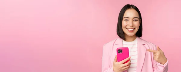 Gülümseyen Asyalı iş kadınının portresi cep telefonuna işaret ediyor, akıllı telefon uygulaması tavsiye ediyor, cep telefonunda başvuru yapıyor, pembe arka planda duruyor. — Stok fotoğraf
