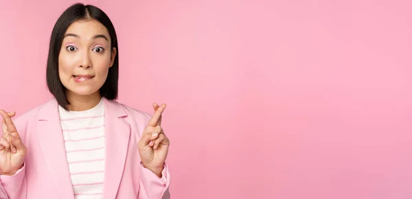 Ευτυχισμένος τυχερός επιχειρηματίας, ασιατική εταιρική κυρία επιθυμία, κάνει ευχή, ελπίζοντας για smth και προσεύχεται, στέκεται στο κοστούμι πάνω από ροζ φόντο — Φωτογραφία Αρχείου
