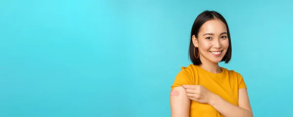 Campaña de vacunas de covid-19. Joven hermosa, saludable mujer asiática mostrando hombro con bandaid, concepto de vacunación, de pie sobre fondo azul — Foto de Stock