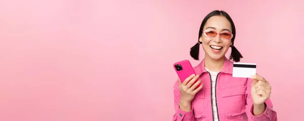 Moderna bella ragazza asiatica, ridendo e sorridendo con il telefono cellulare, carta di credito, shopping online, pagare con smartphone, in piedi su sfondo rosa — Foto Stock