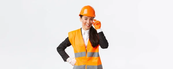 Αυτοπεποίθηση επιτυχημένη ασιατική επικεφαλής διευθυντής αναζητούν υπερήφανοι και χαμογελώντας στην κάμερα, φορώντας κράνος ασφαλείας, γυαλιά και γάντια στο χώρο των κατασκευών ή των επιχειρήσεων, λευκό φόντο — Φωτογραφία Αρχείου