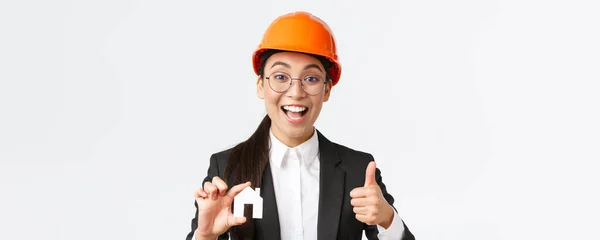 乐观的亚洲女工程师、戴头盔和西装的建筑师的特写镜头鼓励与她的公司联系，以进行翻新和设计，展示房屋和大拇指 — 图库照片