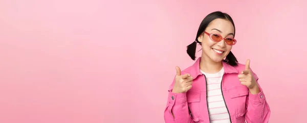 Gros plan portrait de la fille asiatique moderne en lunettes de soleil souriant, pointant du doigt la caméra, vous féliciter, invitant ou complimentant, debout sur fond rose — Photo