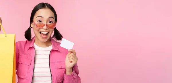 Mutlu genç Asyalı kadın kredi kartı ve alışveriş çantası gösteriyor, mağaza satışı duyurusu yapıyor, dükkanda smth alıyor, pembe arka planda poz veriyor. — Stok fotoğraf