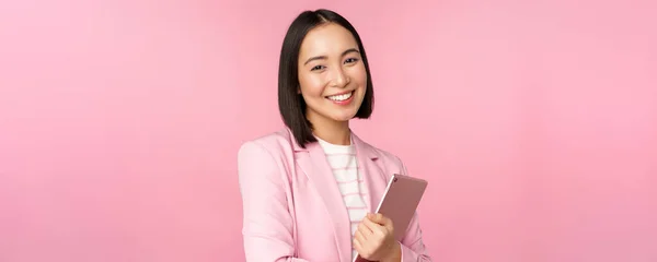 Профессиональная улыбающаяся азиатская бизнесвумен, стоящая с цифровым планшетом, носящая костюм для офисной работы, выглядящая уверенно и счастливо, позирующая на розовом фоне — стоковое фото