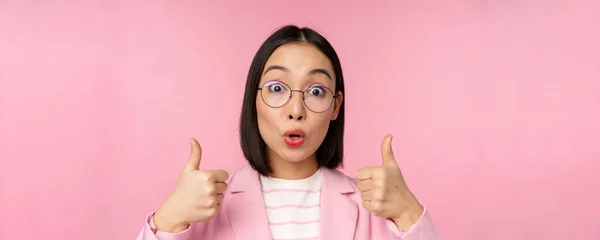 Крупним планом портрет враженої корпоративної жінки, азіатської бізнес-леді в окулярах, показуючи великі пальці вгору, дивлячись здивовано на камеру, рекомендую, рожевий фон — стокове фото