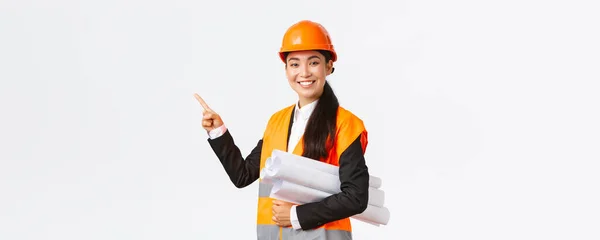 Улыбающаяся профессиональная азиатская женщина-архитектор, инженер в защитном шлеме представляет проект строительства, указывая левой рукой, как носить чертеж, выступая с речью, вводя схему или план строительства — стоковое фото